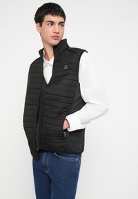 Młody mężczyzna w czarnej pikowanej kamizelce na białej koszuli i niebieskich dżinsach, z rękami w kieszeniach kamizelki, patrzący w bok na białym tle.