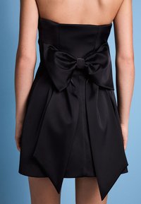 Schwarzes trägerloses Kleid mit einer großen Schleife auf der Rückseite, aus glattem Stoff mit gefalteten Details. Der Rücken ist teilweise freigelegt.