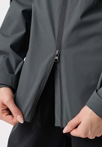 Ruky upravujú alebo rozopínajú spodnú časť tmavosivej bundy s viditeľným zipsom a detailom na manžete.