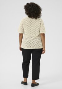 Vrouw met krullend haar, gekleed in een crèmekleurig gebreid T-shirt met korte mouwen en zwarte broek, staande met de rug naar de camera op een effen achtergrond.