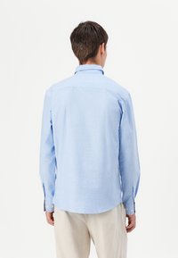 Camisa de botones azul claro, de manga larga, con una tela suave, dobladillo curvado, yugo trasero incorporado, y puños ajustables con botones.