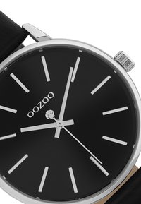 Montre analogique noire avec des index et aiguilles argentés, boîtier rond en acier inoxydable et bracelet en cuir noir. Marquée "OOZOO" sur le cadran.