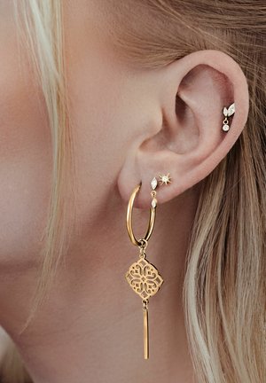 Nahaufnahme eines Ohrs mit blonden Haaren, das vier Ohrringe aus Gold und Diamanten zeigt, darunter einen großen dekorativen Reifen mit einem hängenden Anhänger.