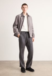 Veste en velours côtelé gris, t-shirt à col rond blanc, pantalons de survêtement gris foncé et chaussures en daim noires. Le mannequin se tient dans un cadre neutre.