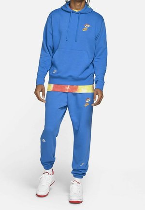 Person trägt blauen Nike-Hoodie und Jogginghose mit weißen und roten Sneakers sowie ein gelb-rotes Batik-Shirt, das unter dem Hoodie hervorblitzt.