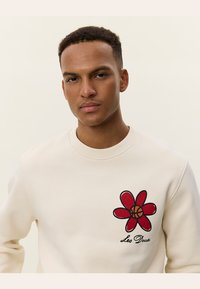 Sudadera color crema con cuello redondo. Presenta un diseño de flor roja con un centro de baloncesto y el texto "Les Deux" en negro.