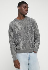 Γκρι πλεκτή ζακέτα με υφασμάτινο σχέδιο, στρογγυλή λαιμόκοψη και ριμπ μανίκια. Συνδυάζεται με ανοιχτού μπλε τζιν παντελόνι.