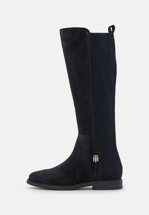 Botte noire mi-mollet avec devant en daim, dos élastique côtelé, talon bas et détail monogramme en métal près de la cheville sur fond uni.