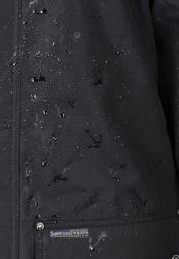 Chaqueta negra impermeable con gotas de agua, que presenta un sutil patrón de anclas y un parche con el logo en la parte inferior izquierda. Textura suave.