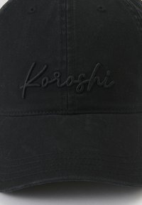 Črna bombažna baseball kapa s ukrivljenim robom in vezenim logotipom "Koroshi" v usklajeni barvi na sprednjem delu. Teksturirana tkanina.