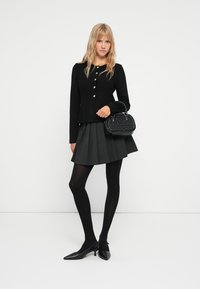 Cardigan noir avec boutons argentés, jupe plissée grise, collants noirs et escarpins noirs pointus. Petit sac à main noir avec un design texturé.