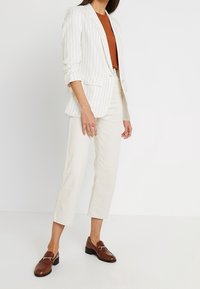 Blazer blanc à rayures avec une coupe ajustée, porté sur un haut marron. Associé à un pantalon court crème et des mocassins marron, avec une texture lisse.