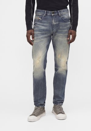 Hombre vistiendo vaqueros azules desgastados y descoloridos con pequeños desgarros en los muslos, combinados con zapatillas altas grises y una camiseta negra de manga larga.