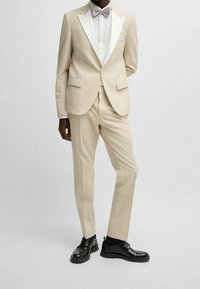 Uomo in abito beige con rever bianchi, camicia bianca, papillon beige e scarpe eleganti nere, in piedi contro uno sfondo semplice.