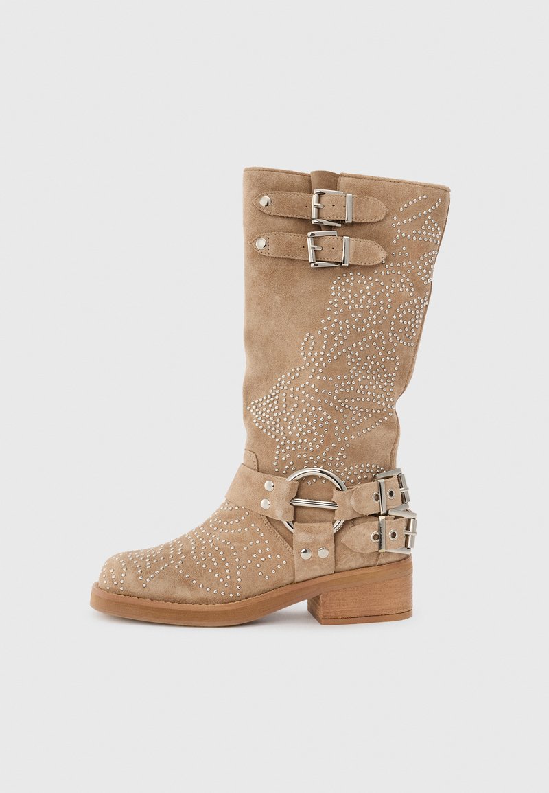 Bota de ante color marrón claro, hasta la rodilla, con remaches decorativos de plata, acentos de doble hebilla, punta redondeada y un tacón de madera grueso.