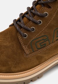 Brown suede boot with dark brown laces, metal eyelets, and green embroidered logo. Features a textured sole and contrasting beige midsole.