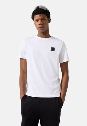 Ung mand iført hvid T-shirt med korte ærmer og lille sort logoemblem, sorte bukser og solbriller hvilende på hans hoved.