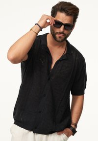 Schwarzes, kurzärmliges Strickshirt mit strukturiertem Muster und Knopfverschluss. Modell trägt Sonnenbrille und präsentiert das offene Design des Kleidungsstücks.