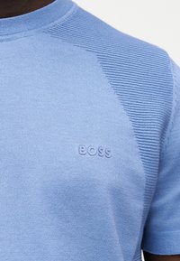 Ανοιχτό μπλε πλεκτό πουκάμισο με κοντά μανίκια, υφασμένα πάνελ στους ώμους και ανάγλυφο λογότυπο BOSS στο στήθος, φορεμένο από άτομο.