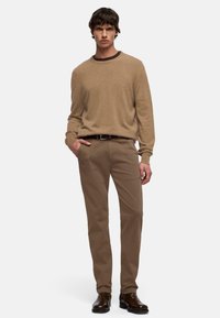 Beige gebreide trui met een ronde hals, gecombineerd met beige chino's en bruine leren laarzen. Model staat met één hand in de zak.