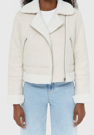 Femme portant une veste en peau retournée couleur crème avec une fermeture éclair asymétrique, une chemise blanche et un jean taille haute bleu clair.