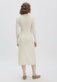 Abito a maglia a maniche lunghe di un beige chiaro, con vita aderente e design con cintura, che arriva a metà polpaccio e presenta una texture a costine. Scarpe piatte visibili.