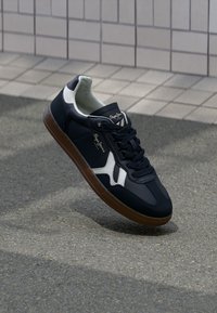 Mørkeblå sneakers med hvite detaljer og logo, teksturert overdel, snøring, kontrastfylt brun gummisåle, glatte og holdbare materialer.