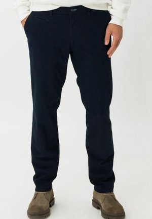 Pantaloni di cotone blu navy con vestibilità slim, tasche frontali e zip. Abbinati a stivali alla caviglia marroni, che mostrano un design pulito e una texture sottile.