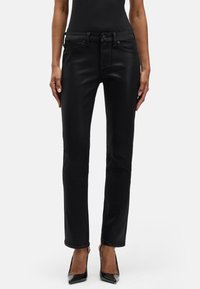 Kobieta ubrana w błyszczące, czarne, obcisłe dżinsy i czarne buty na wysokim obcasie, stojąca na tle jednolitego, białego tła.