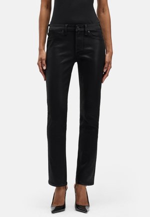 Frau trägt glänzende schwarze Slim-Fit-Jeans und schwarze High-Heel-Schuhe, steht vor einem schlichten weißen Hintergrund.