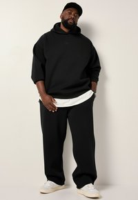 Zwarte oversized hoodie met een logo aan de voorkant, gecombineerd met zwarte brede broek. De outfit heeft een witte gelaagde onderhemd en witte sneakers.