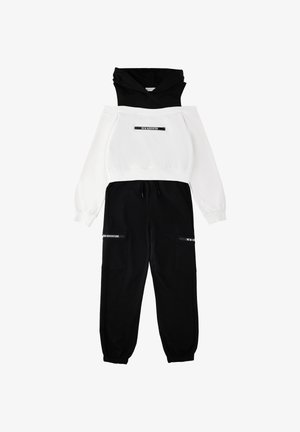 Felpa con cappuccio bianca e nera con la scritta "NEW ADVENTURE", abbinata a pantaloni cargo neri con la stessa scritta sulle tasche.