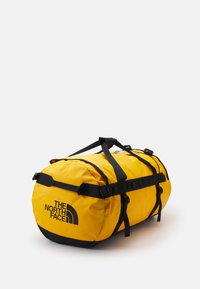 Žuta torba s remenima i detaljima u crnoj boji. Ima zaobljen oblik i logo "THE NORTH FACE" u crnoj boji. Teksturirana površina tkanine.