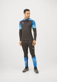 Puma-Sportoutfit mit schwarzem Oberteil und Ärmeln mit blauem Farbverlauf, mit einem Halb-Reißverschluss-Kragen, schlanker Passform und strukturierten Akzenten an den Beinen.