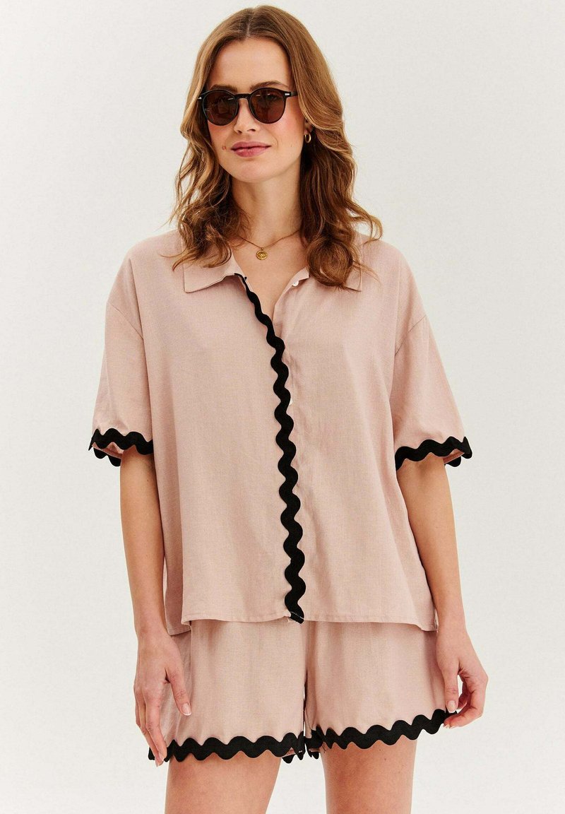 Svetloružová košeľa s krátkym rukávom a čiernym rickrack lemom pozdĺž goliera a rukávov, doplnená o zladené ružové šortky s podobným lemovaním.