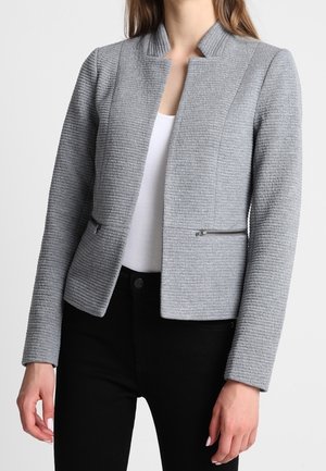 Kvinde iført en tætsiddende lysegrå tekstureret blazer med lynlåslommer over en hvid top og sorte bukser.