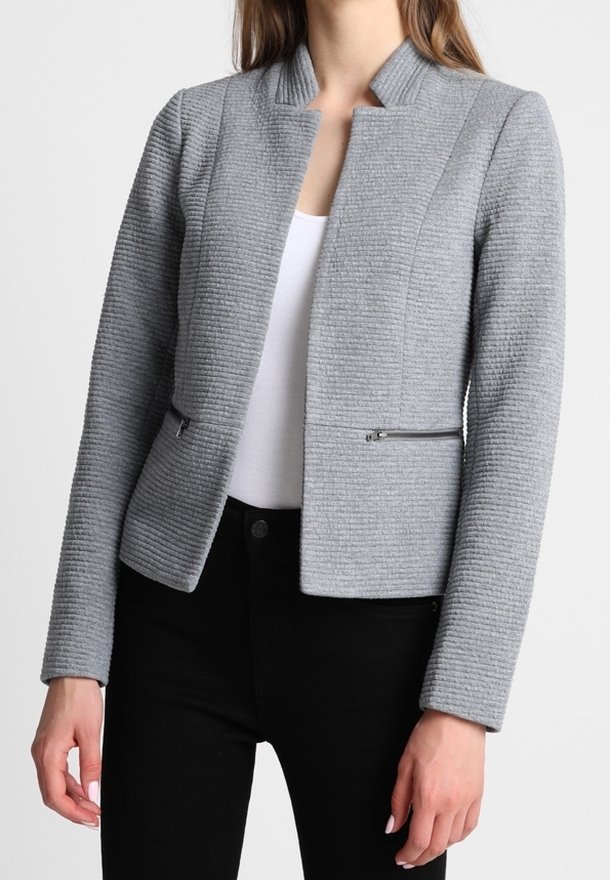 Femme portant un blazer ajusté gris clair texturé avec des poches zippées sur un haut blanc et un pantalon noir.