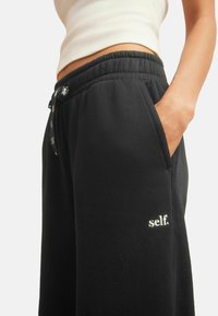 Person, der schwarze Jogginghosen trägt, mit "self." auf der Nähe der Tasche bestickt, Hand in der Tasche ruhend, cropped weißes ärmelloses Oberteil sichtbar.