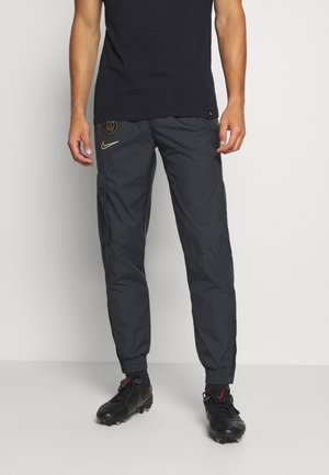 Mężczyzna w czarnych sportowych spodniach Nike z logo PSG i czarnych korkach, stojący na tle jednolitego białego tła.