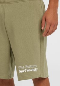Olijfgroene shorts van zacht materiaal, met zijzakken en een witte bedrukte logo op het linker onderbeen.