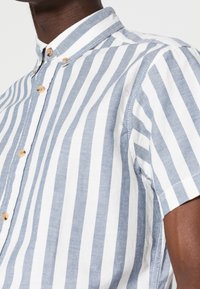 Kortärmad skjorta med knappar, prydd med växlande blå och vita vertikala ränder, träknappar och en klassisk krage.