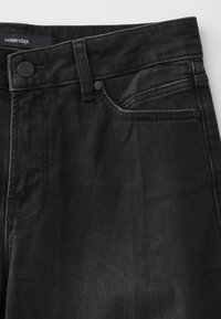 Gros plan sur un jean denim noir montrant la ceinture, le bouton, la boucle de ceinture, la poche avant et l’étiquette de la marque indiquant "someday."