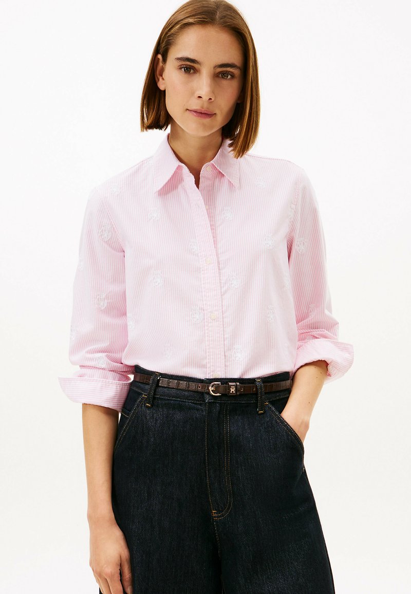 Light pink striped button-up shirt with white floral embroidery, rolled sleeves, and a black belt worn over dark denim jeans.
