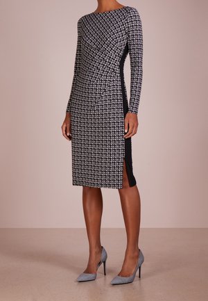 Femme portant une robe noire et blanche à carreaux pied-de-poule, à manches longues, longueur genou, avec une fente latérale, et des chaussures à talons assorties.