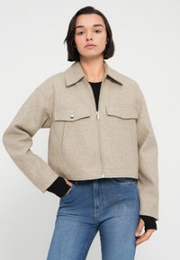 Μπεζ cropped μπουφάν με γιακά, κλείσιμο με φερμουάρ και δύο τσέπες στο στήθος. Το υλικό φαίνεται μαλακό με υφή.