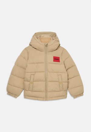 Beige polstret hettejakke med glidelås, to frontlommer med knapp, elastiske mansjetter, og rød rektangulær lapp merket "HUGO" på brystet.