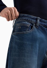 Jeans de mezclilla azul con un ajuste slim, con costuras de los bolsillos traseros en dorado y un pequeño logo de A/X. La tela muestra un aspecto desgastado.
