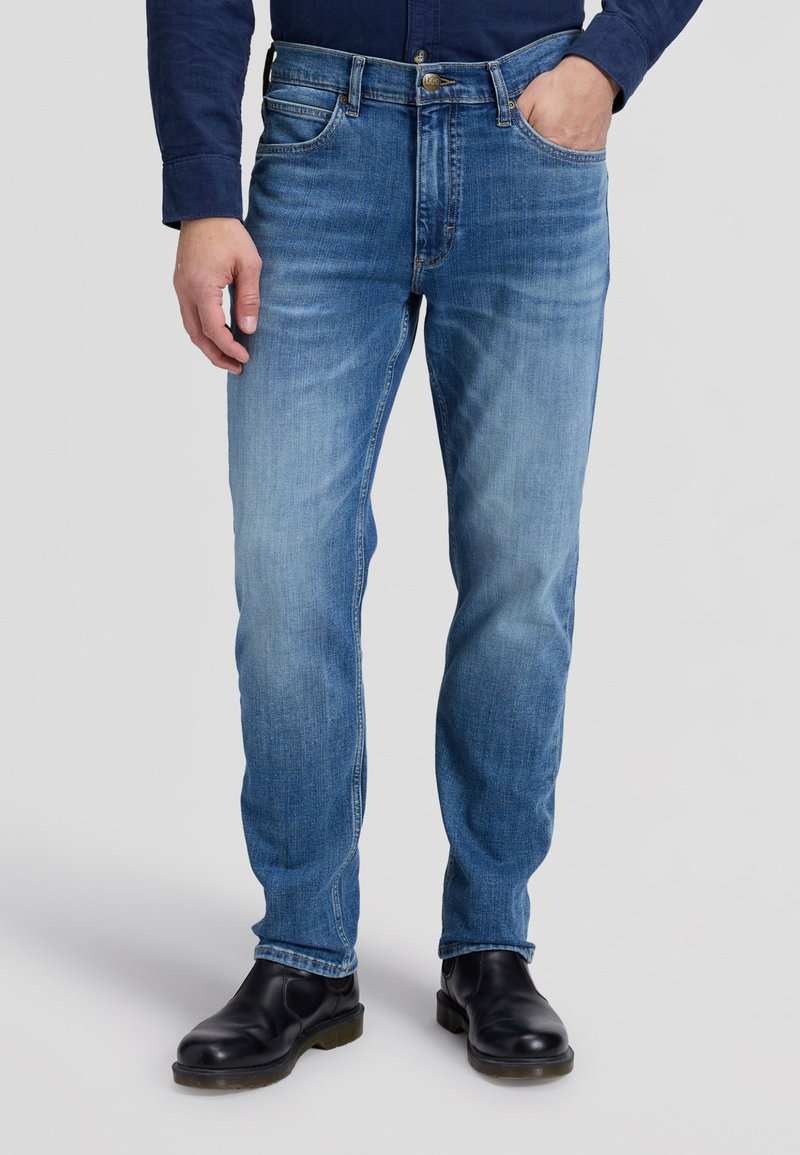 Homme portant un jean bleu délavé droit, des bottes en cuir noires, et une chemise bleu marine à manches longues avec une main dans la poche.