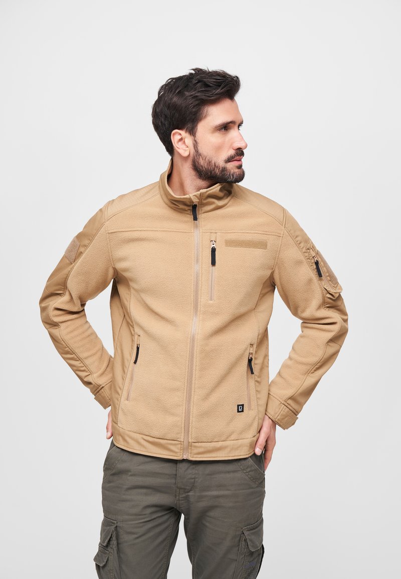 Giacca in pile beige con colletto alto, tasche con zip e toppe sulle spalle. Caratterizzata da un tessuto testurizzato e un design aderente.
