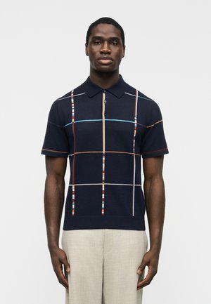 Uomo che indossa una polo blu navy a maniche corte con strisce verticali e orizzontali multicolori, abbinata a pantaloni beige chiaro.